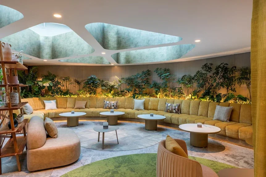 A curved lounge with green velvet banquettes, indoor plants and sculpted skylights